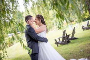 Un couple partageant un moment tendre sous le feuillage d'un saule pleureur. La mariée, vêtue d'une robe de mariage blanche fluide, touche doucement le visage du marié alors qu'ils se regardent dans les yeux. Le marié est habillé d'un costume noir classique. En arrière-plan, on peut voir une pelouse verte luxuriante avec plusieurs chaises Adirondack en bois, suggérant un cadre de mariage en plein air. La scène est sereine et romantique, encadrée par la verdure douce de l'arbre.